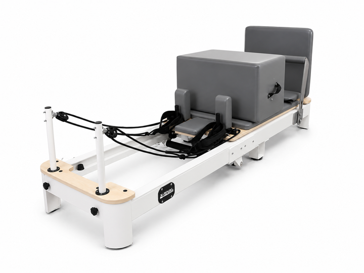 A Pilates reformer machine with a white frame, natural wood accents, black straps, and a gray padded carriage and backrest, shown on a clean white background.